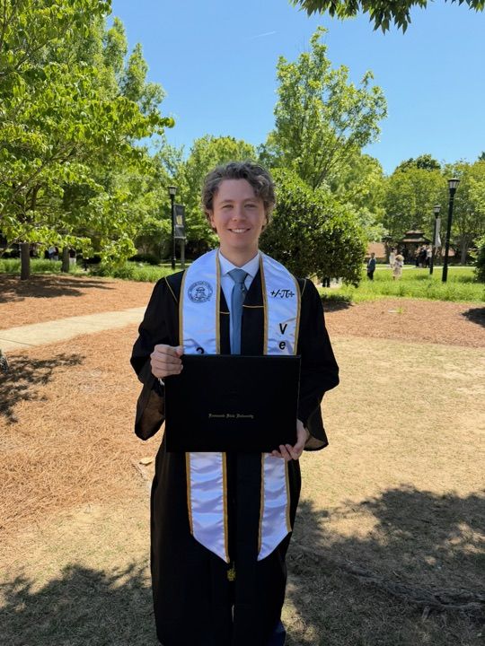 Grant Versluis at his university graduation, holding his diploma, in a cap and gown with a stole for a service fraternity